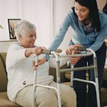 A home health aide helping an elderly woman get up from the couch using a walker.