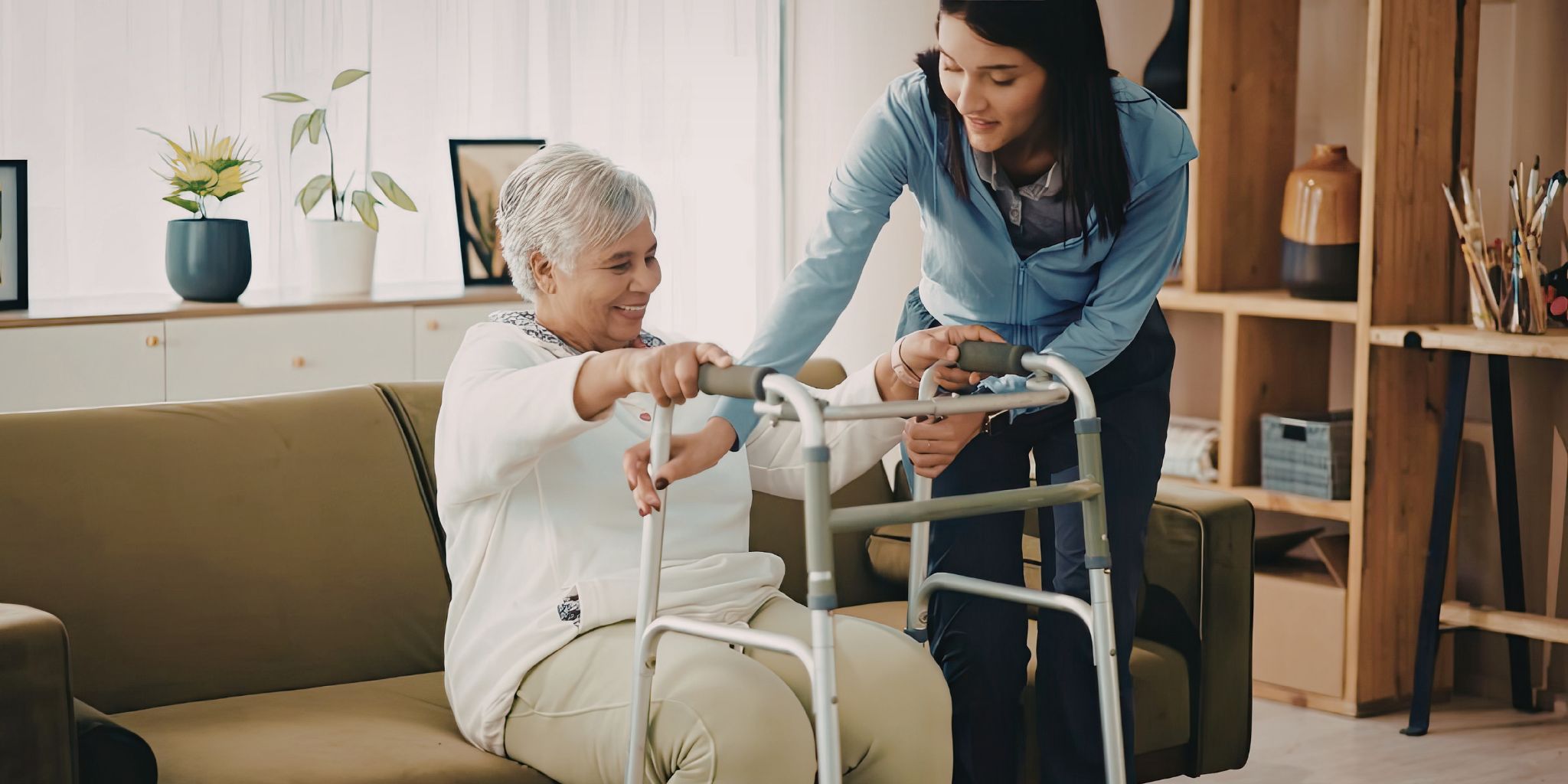 A home health aide helping an elderly woman get up from the couch using a walker.
