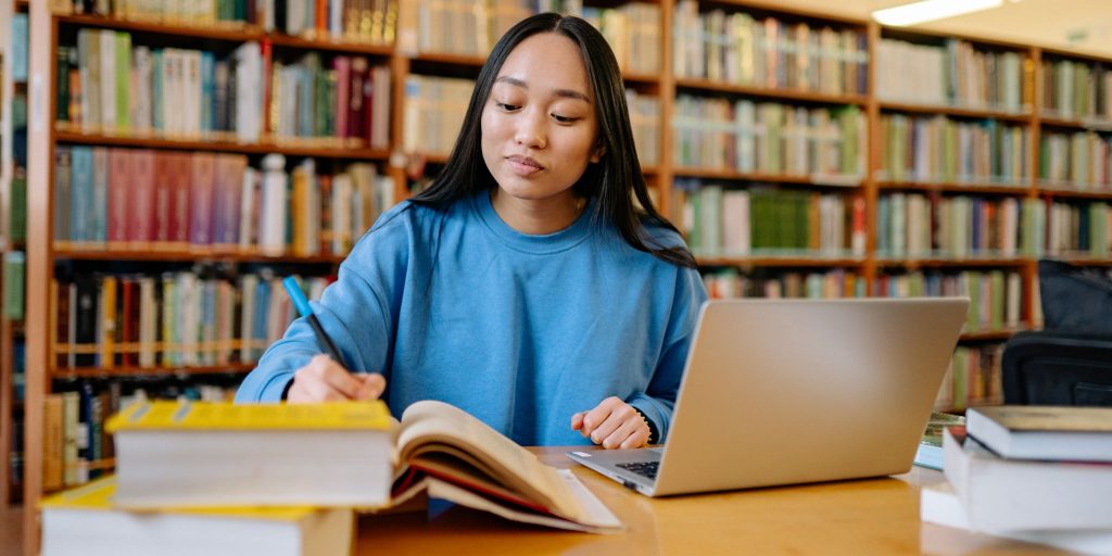 College student in library working at a desk with books and writing in a notebook in front of a laptop.
