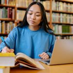 College student in library working at a desk with books and writing in a notebook in front of a laptop.