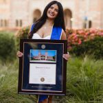 Smiling woman outdoors in front of ornamental grasses and pink flowers holding a UCLA diploma frame with a photo of Royce Hall building