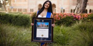 Smiling woman outdoors in front of ornamental grasses and pink flowers holding a UCLA diploma frame with a photo of Royce Hall building