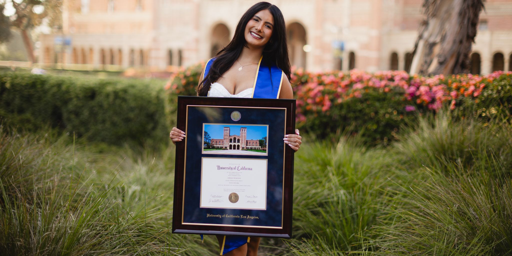Smiling woman outdoors in front of ornamental grasses and pink flowers holding a UCLA diploma frame with a photo of Royce Hall building