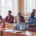Classroom of young adults smiling and listening to a professor at front of class.