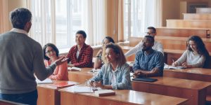 Classroom of young adults smiling and listening to a professor at front of class.