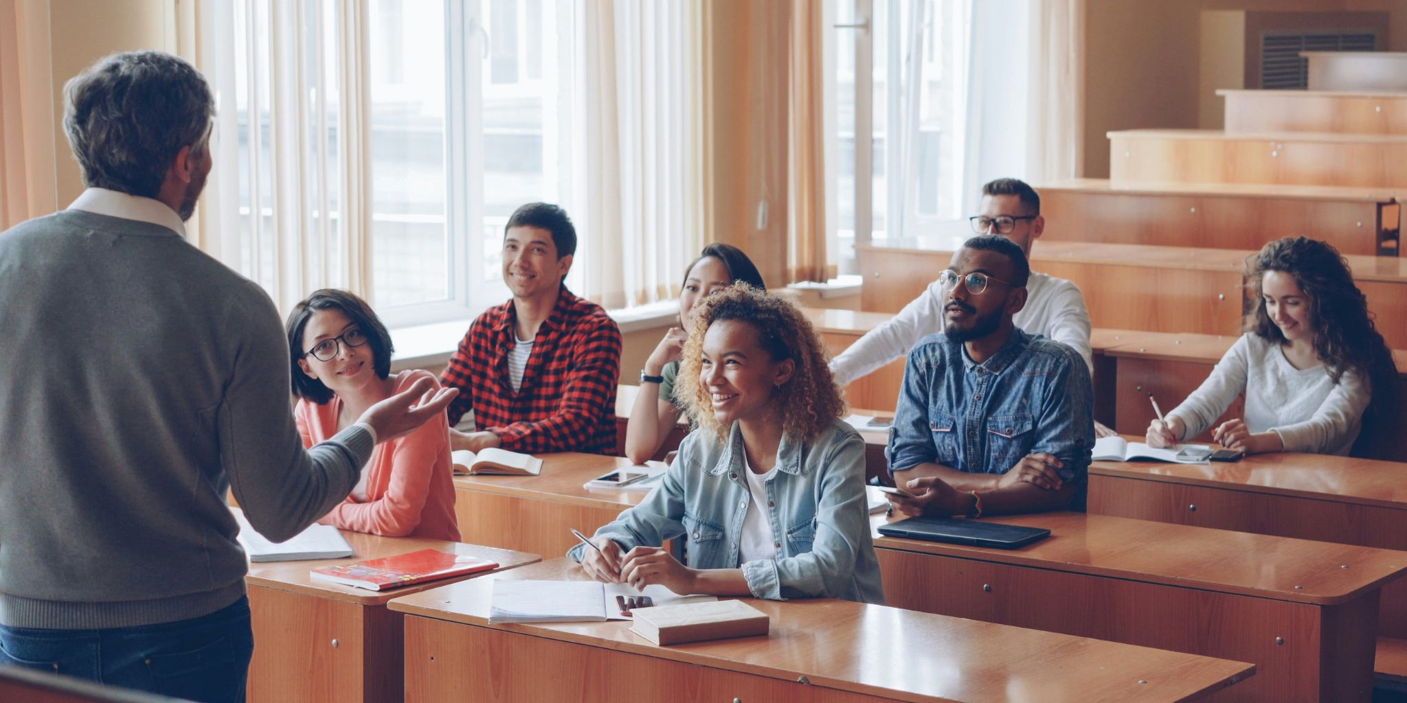 Classroom of young adults smiling and listening to a professor at front of class.