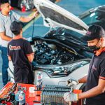 People working in an automative garage on a car with hood up.