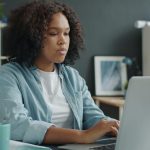 College student sitting at desk working on laptop with aqua coffee next to her and bookshelves behind her.