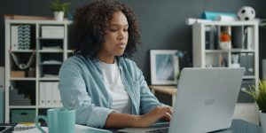 College student sitting at desk working on laptop with aqua coffee next to her and bookshelves behind her.