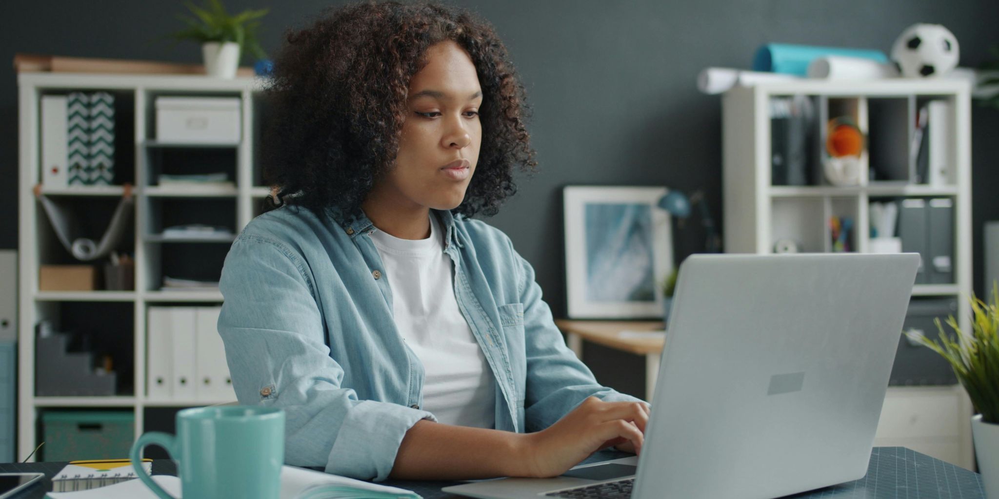 College student sitting at desk working on laptop with aqua coffee next to her and bookshelves behind her.