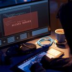 A darkly lit photo showing a cybercriminal in a hoodie working on a laptop with a screen that says "Access Denied".