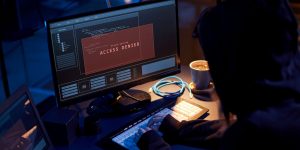 A darkly lit photo showing a cybercriminal in a hoodie working on a laptop with a screen that says "Access Denied".