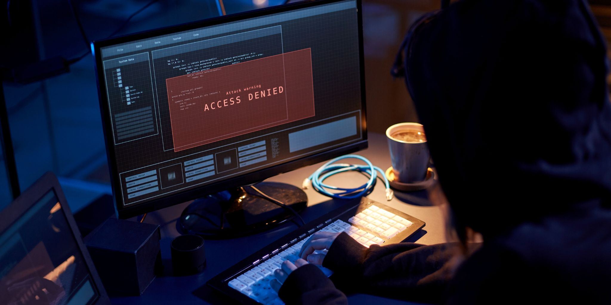 A darkly lit photo showing a cybercriminal in a hoodie working on a laptop with a screen that says "Access Denied".