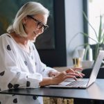 Smiling woman in glasses working on her laptop in a well-lit office. She's wearing a blousy white shirt with large black dots.