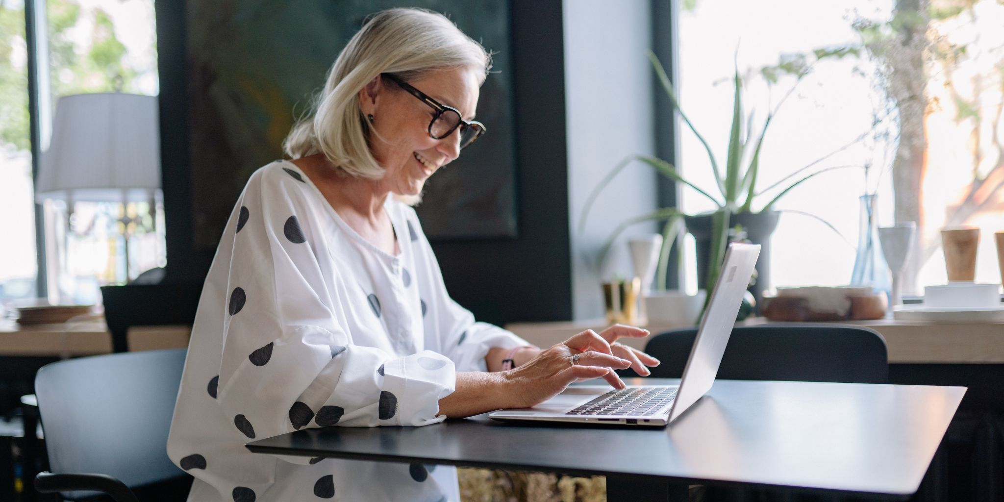 Smiling woman in glasses working on her laptop in a well-lit office. She's wearing a blousy white shirt with large black dots.