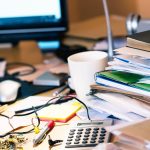 Messy desk with stacks of papers, books, glasses, mug, and more.