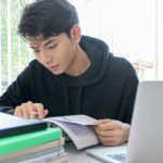 Student in hoodie sitting at desk with pile of books, laptop, studying a textbook with a pencil in his hand.