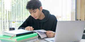 Student in hoodie sitting at desk with pile of books, laptop, studying a textbook with a pencil in his hand.