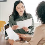 Two women in bright office space with one interviewing the other, both holding papers and talking.