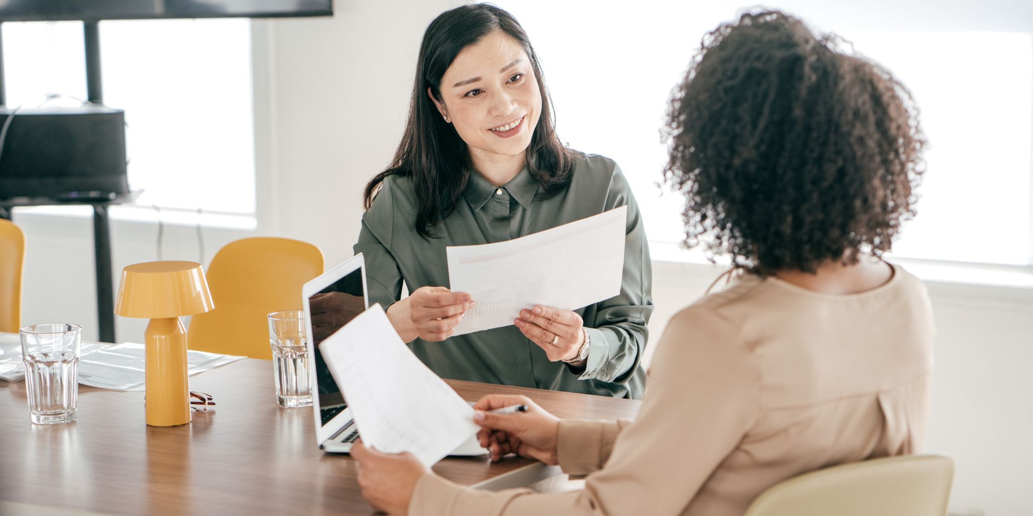 Two women in bright office space with one interviewing the other, both holding papers and talking.