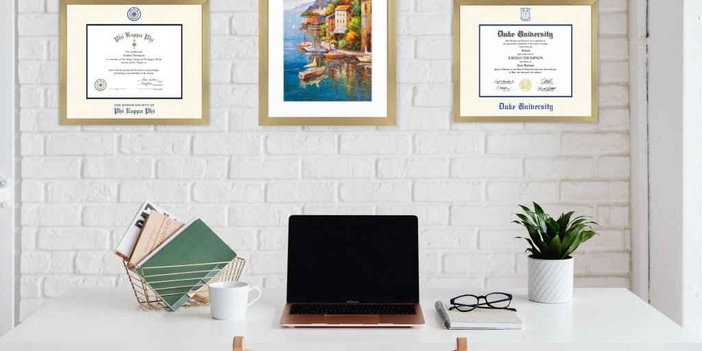 Home office with white brick wall with a Phi Kappa Phi certificate frame, colorful poster of coastal village, and a Duke University diploma frame, all in gold frames.