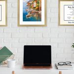 Home office with white brick wall with a Phi Kappa Phi certificate frame, colorful poster of coastal village, and a Duke University diploma frame, all in gold frames.
