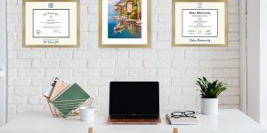 Home office with white brick wall with a Phi Kappa Phi certificate frame, colorful poster of coastal village, and a Duke University diploma frame, all in gold frames.