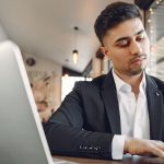 Man in suit working at desk writing in notebook with pen next to a silver laptop