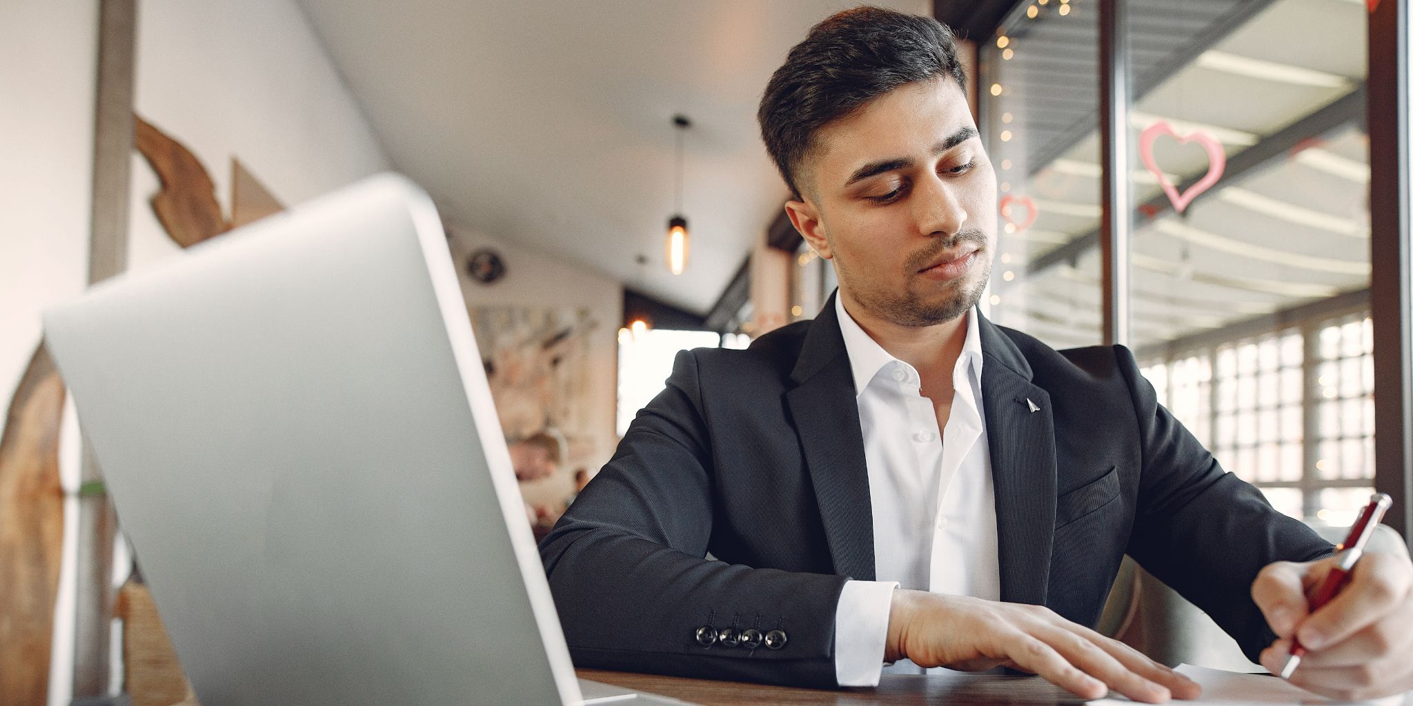 Man in suit working at desk writing in notebook with pen next to a silver laptop