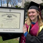 Smiliing grad in cap and gown outdoors holding a Fordham University diploma frame.