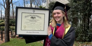 Smiliing grad in cap and gown outdoors holding a Fordham University diploma frame.