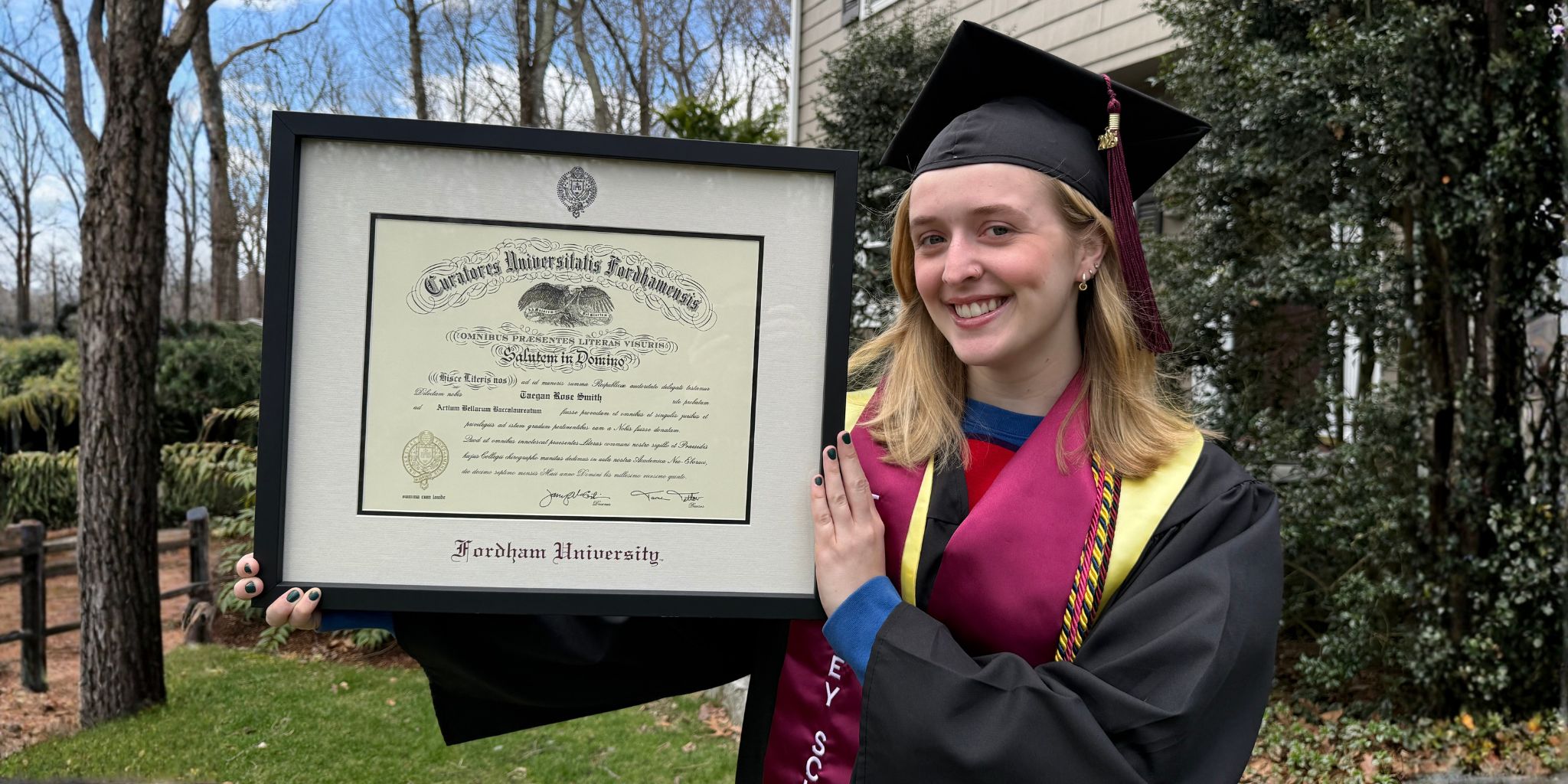 Smiliing grad in cap and gown outdoors holding a Fordham University diploma frame.