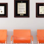 Waiting room with three orange chairs, framed Hippocratic Oath, and two medical degree frames on wall with pink lamp to one side and colorful houseplant on table on other side of chairs.