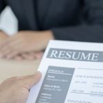 Closeup of the hands of an interviewer holding a paper that says "Resume" and lists job experiences while a job applicant sits across from them at a desk.