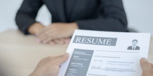 Closeup of the hands of an interviewer holding a paper that says "Resume" and lists job experiences while a job applicant sits across from them at a desk.