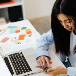Woman at her desk working on her phone with her laptop, notes, and color swatches nearby.
