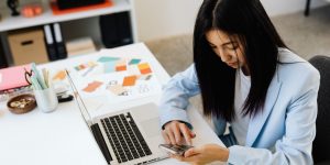 Woman at her desk working on her phone with her laptop, notes, and color swatches nearby.
