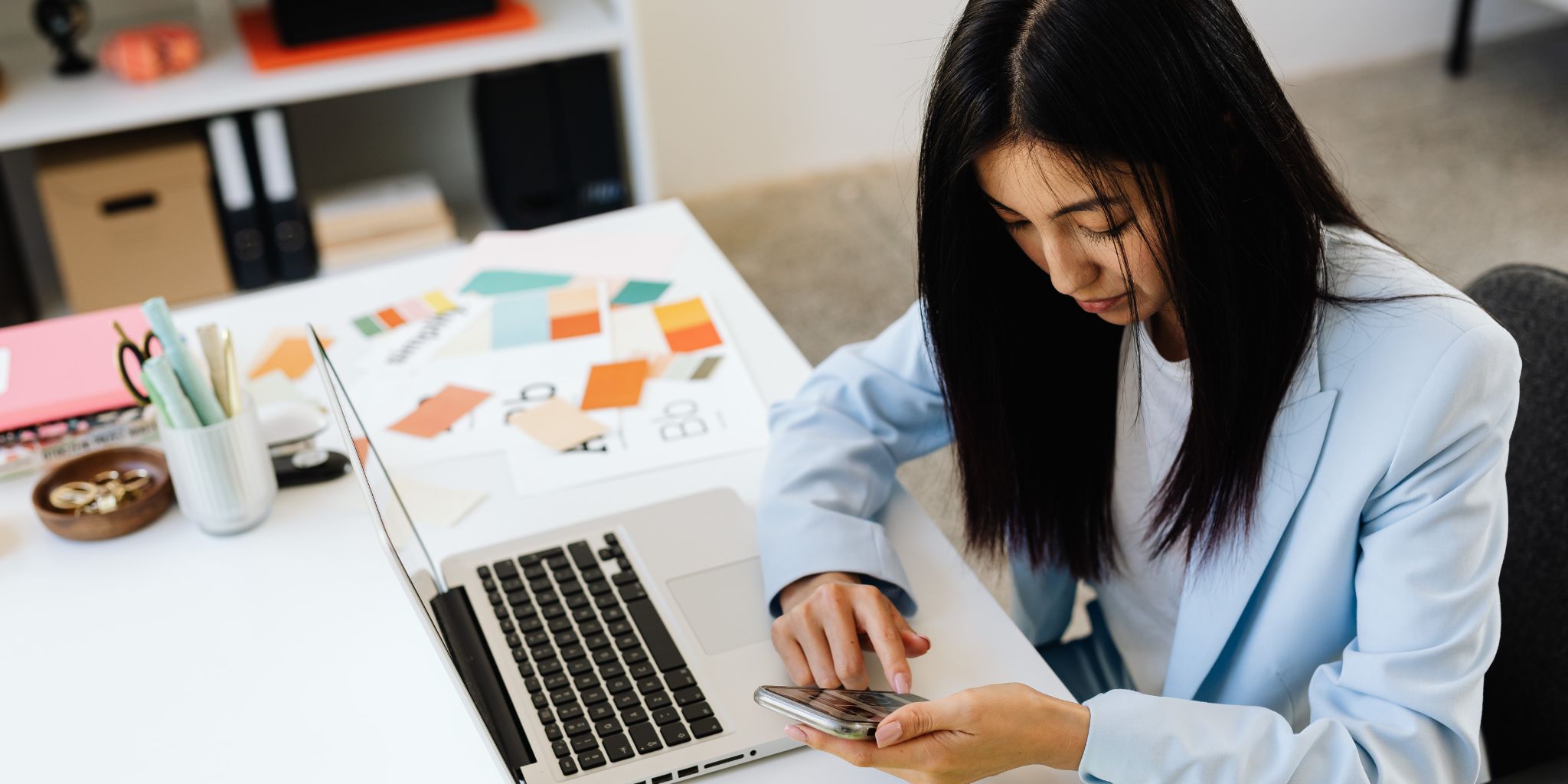 Woman at her desk working on her phone with her laptop, notes, and color swatches nearby.