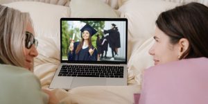 Closeup of mother and daughter looking at each other, working on a bed on a laptop with a photo of girl in graduation cap and gown holding a degree with a red ribbon.