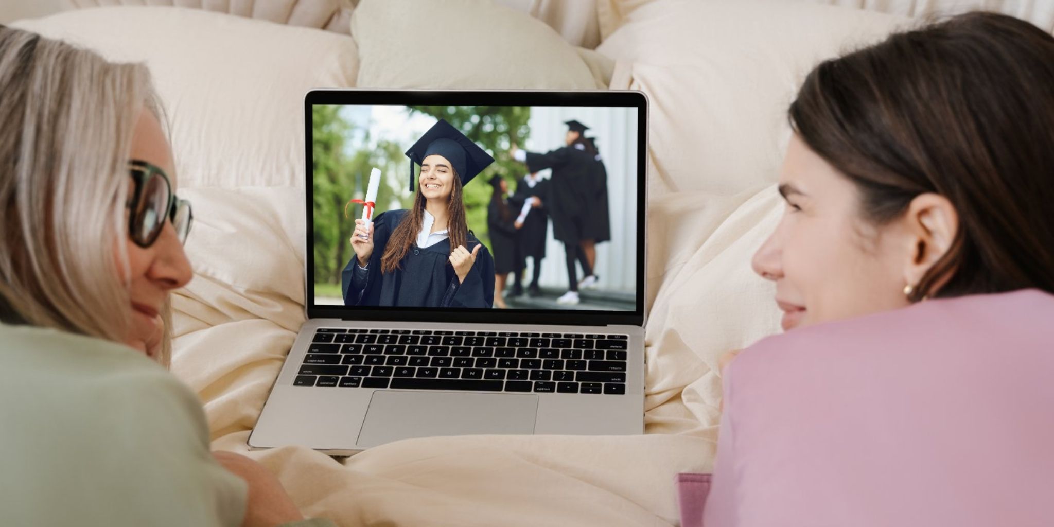 Closeup of mother and daughter looking at each other, working on a bed on a laptop with a photo of girl in graduation cap and gown holding a degree with a red ribbon.