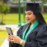 Graduate in cap and gown with green graduation sash on phone typing outside on campus.
