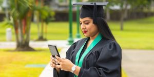 Graduate in cap and gown with green graduation sash on phone typing outside on campus.