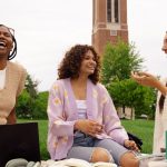 Three college students sitting on grass on college campus studying with large tower behind them.