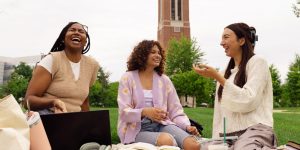Three college students sitting on grass on college campus studying with large tower behind them.