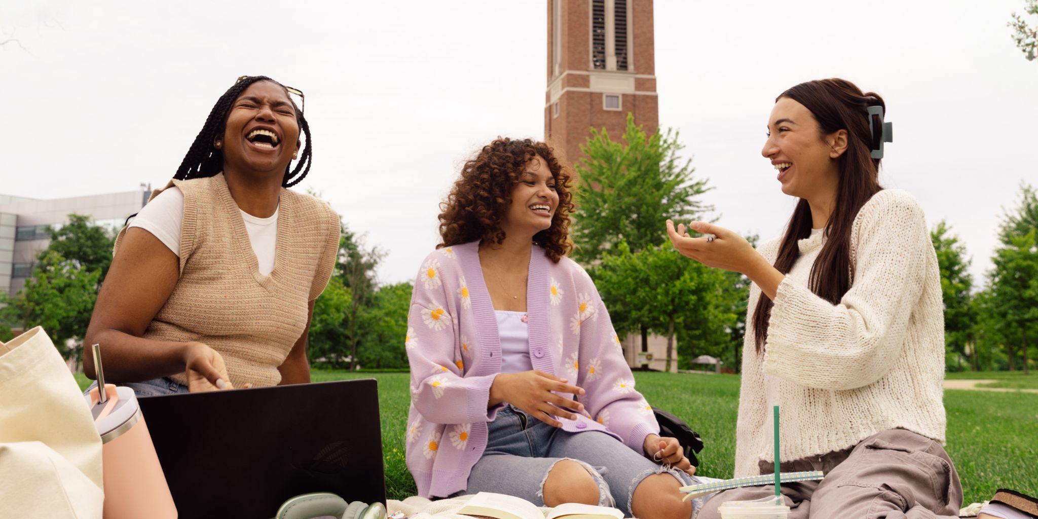 Three college students sitting on grass on college campus studying with large tower behind them.