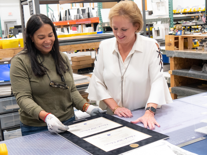 Two women, one is Lucie Voves CEO of Church Hill Classics, in a workshop carefully inspecting a handcrafted diploma frame at their production facility surrounded by tools and framing materials