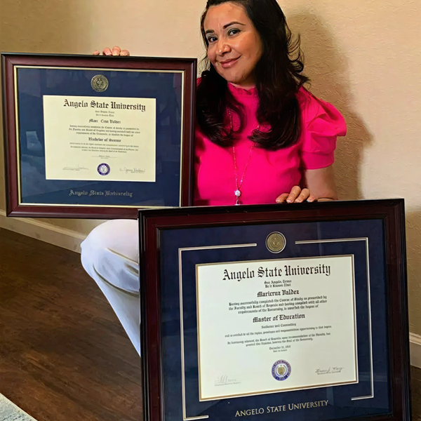 Woman in a bright pink blouse holding two framed Angelo State University diplomas with dark wood frames and blue matting.
