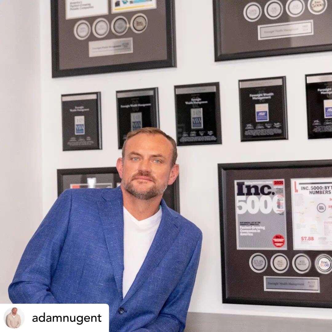 Man in a blue blazer standing in front of a wall filled with framed business awards, plaques, and Inc. 5000 recognitions.