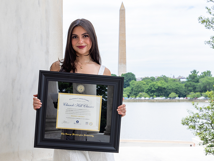 Young woman holding a diploma frame with the Washington Monument in the background, smiling at the camera.