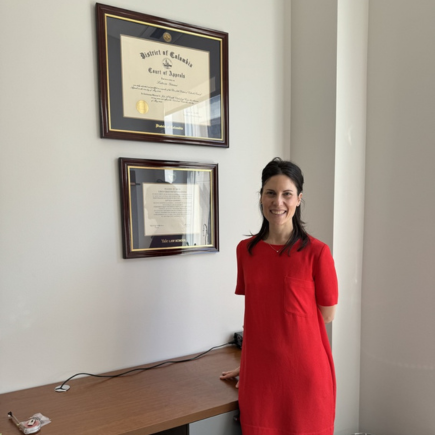 Smiling woman in a red dress standing beside a desk, beneath two professionally framed diplomas mounted on a white wall.